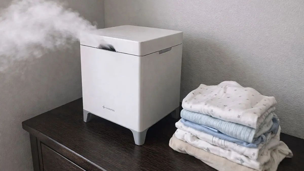 Carepod Cube Plus X50V, the best cool mist humidifier for baby nurseries, sitting on a wooden dresser next to folded baby clothes