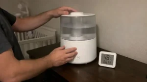 A dad setting up a modern white cool-mist humidifier on a dark wood nursery dresser next to a digital hygrometer reading 48% humidity.