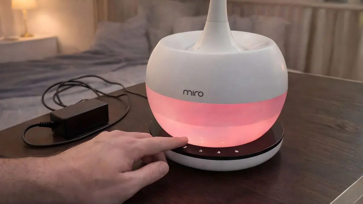A medium shot focusing on the fully assembled white Miro NR08M humidifier on a nursery dresser with a dad's hand hovering above the separate Luma Touch external control panel